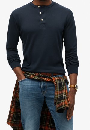 Man draagt een marineblauw henley-shirt met lange mouwen, blauwe spijkerbroek, geruit overhemd om de taille geknoopt en een gouden horloge aan zijn linkerpols.