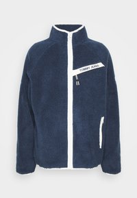Námorně modrý fleecový kabát s bílým zipem a prvky. Obsahuje nášivku na hrudi a boční kapsy, strukturovanou tkaninu a vysoký límec.
