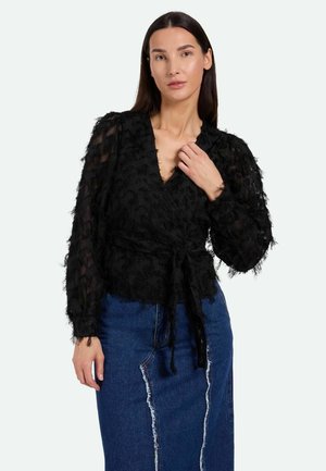 Blusa nera avvolgente in tessuto testurizzato, con profondo scollo a V e lunghe maniche trasparenti; legata in vita. Abbinata a una gonna di jeans blu con dettagli consumati.