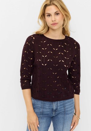 Lyshåret kvinde iført en mørkelilla strikket sweater med bladformede udskæringer og blå jeans, stående med den ene hånd ned langs siden.