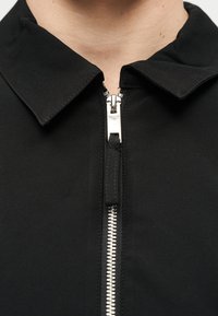 Κοντινό πλάνο ενός μαύρου μπουφάν με ασημί μεταλλικό φερμουάρ εν μέρει κλειστό και διπλωμένο γιακά, που φοριέται στον λαιμό και το πάνω μέρος του στήθους ενός ατόμου.