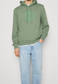 Man i salviafärgad huvtröja med framlogo och ljusblå jeans, står med ena handen i fickan mot en enkel bakgrund.