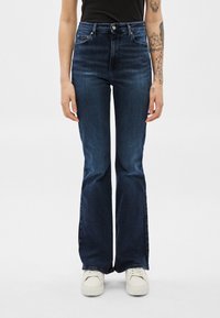 Cilvēks, kurš valkā tumši zilas augstās vidukļa zvana bikses, baltas sporta kurpes un melnu krekliņu, stāv taisni ar redzamām tetovējumu zīmēm uz labās rokas.