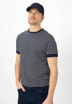 Mann mit marineblauer Mütze, der ein marineblau-weiß gestreiftes Kurzarmhemd trägt, mit den Händen in den Taschen dunkler Jeans, blickt leicht nach oben vor weißem Hintergrund.