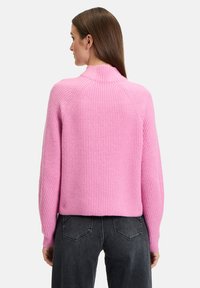 Rosa geribbelde trui met een hoge nek en raglanmouwen. Heeft een textuurpatroon. G gedragen met donkergrijze jeans.