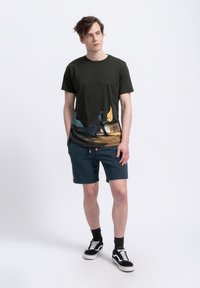 T-shirt in cotone verde scuro con una stampa grafica colorata nella parte inferiore; abbinato a pantaloni corti turchesi e sneakers nere e bianche.
