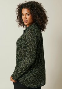 Camisa de manga larga de color verde oscuro con un patrón zigzag abstracto en negro, cuello abotonado y corte relajado. Textura de tela suave con aberturas laterales.