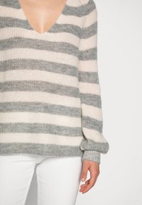 Sweter w paski z dekoltem w kształcie litery V, w jasnoróżowe i szare paski, z prążkowanymi mankietami oraz miękką, teksturowaną dzianiną. Połączony z białymi spodniami.