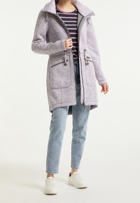 Žena v světle fialové zipové bundě s kapucí, pruhovaném tričku, světle modrých džínách a bílých teniskách, stojící na jednobarevném pozadí.