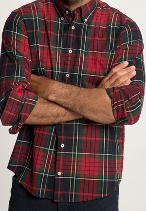Mężczyzna ze skrzyżowanymi ramionami, ubrany w czerwoną, zieloną i czarną kraciastą koszulę zapinaną na guziki, z podwiniętymi rękawami na jednolitym tle.