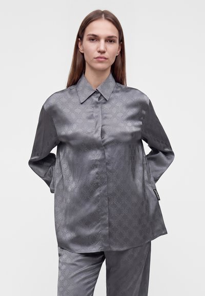 Femme portant une chemise ample en satin gris et un pantalon assorti avec une texture subtilement imprimée, debout devant un fond blanc uni.