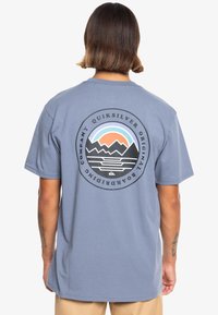 Light blue T-shirt with short sleeves featuring a circular mountain and sun graphic in black and pastel colors on the back.