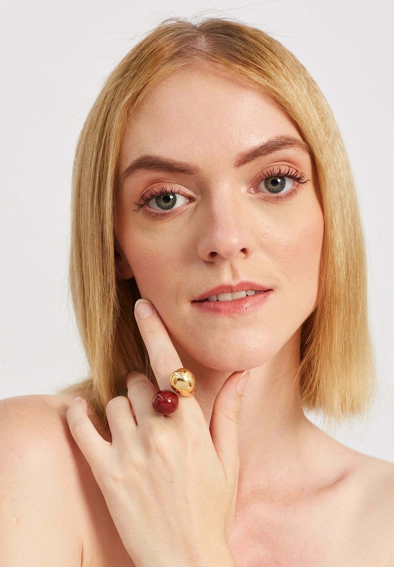 Złote i czerwone pierścionki na nagiej dłoni. Złoty pierścionek ma odbijającą powierzchnię, podczas gdy czerwony pierścionek jest gładki i błyszczący.