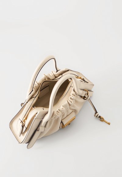 Petit sac à main en cuir beige avec fermeture éclair dorée et breloque clé, côtés doux froncés et deux poignées, présenté ouvert sur une surface blanche.