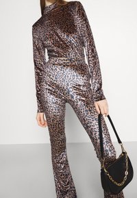 Jumpsuit in tessuto brillante con stampa leopardata bronzo, caratterizzato da maniche lunghe e collo alto, abbinato a una piccola borsa nera con dettagli in catena.