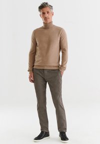 Beżowy sweter z golfem wykonany z miękkiego materiału, zestawiony z teksturowanymi, ciemnobrązowymi spodniami i czarnymi sneakersami, stojący na jednolitym tle.