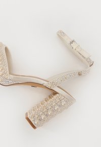 Beige augstpapēžu sānītes ar rotātu bloku papēdi, izrotātas ar pērļu un rhinestone detaļām; ar siksnu un sprādzes aizdari.