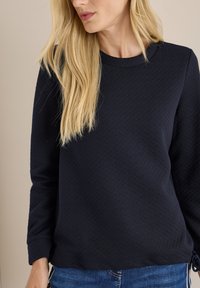 Navyblauwe, gestructureerde sweatshirt met lange mouwen, ronde halslijn en geplooide accenten bij de manchetten. Gepaard met blauwe spijkerbroeken.