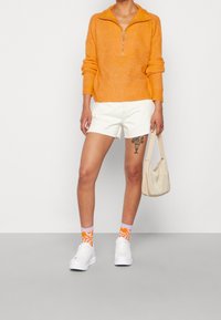 Person wearing an orange zip-up sweater, white shorts, white sneakers, orange patterned socks, holding a beige handbag with a floral tattoo on left thigh.