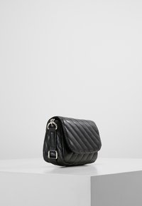 Borsa crossbody in pelle nera trapuntata con chiusura a pattina e finiture in argento su un piedistallo bianco contro uno sfondo grigio.