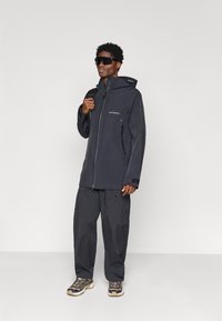 Muž v čiernej vonkajšej bunde a nohaviciach, s veľkými čiernymi slnečnými okuliarmi, nesúci ramenný popruh od batohu, stojaci na čistom bielom pozadí.