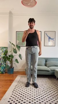 Hombre que lleva una camiseta sin mangas negra, jeans de un azul claro y zapatos negros, de pie sobre una alfombra con patrón, con una planta verde y un sofá en el fondo.