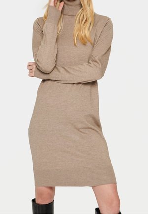 Femme portant une robe-pull à col roulé beige à manches longues et des bottes noires montantes, debout les bras croisés et la main près du visage.