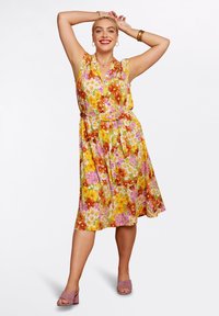 Blommig klänning med livfulla mönster i gult, rosa och orange. Korta ärmar, midjeband och knälång design, kombinerad med mauve skor.