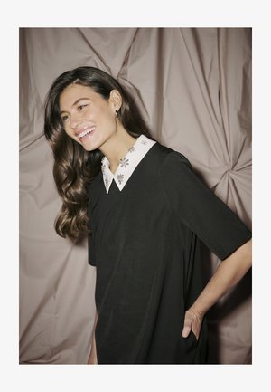 Zwarte jurk met korte mouwen en een witte kraag met bloemige versieringen. Gladde textuur met een ontspannen pasvorm.