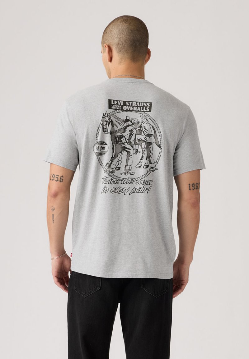 T-shirt gris en coton avec un imprimé graphique noir au dos, comportant des éléments de design vintage et du texte. Manches courtes et coupe décontractée.