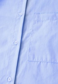 Camicia button-up blu chiaro con righe verticali, caratterizzata da una tasca sul petto e bottoni bianchi realizzati in tessuto liscio e intrecciato.