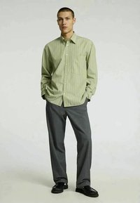 Camisa de botão verde às riscas com um bolso no peito, acompanhada de calças cinzentas. O conjunto apresenta um ajuste descontraído e uma textura suave.