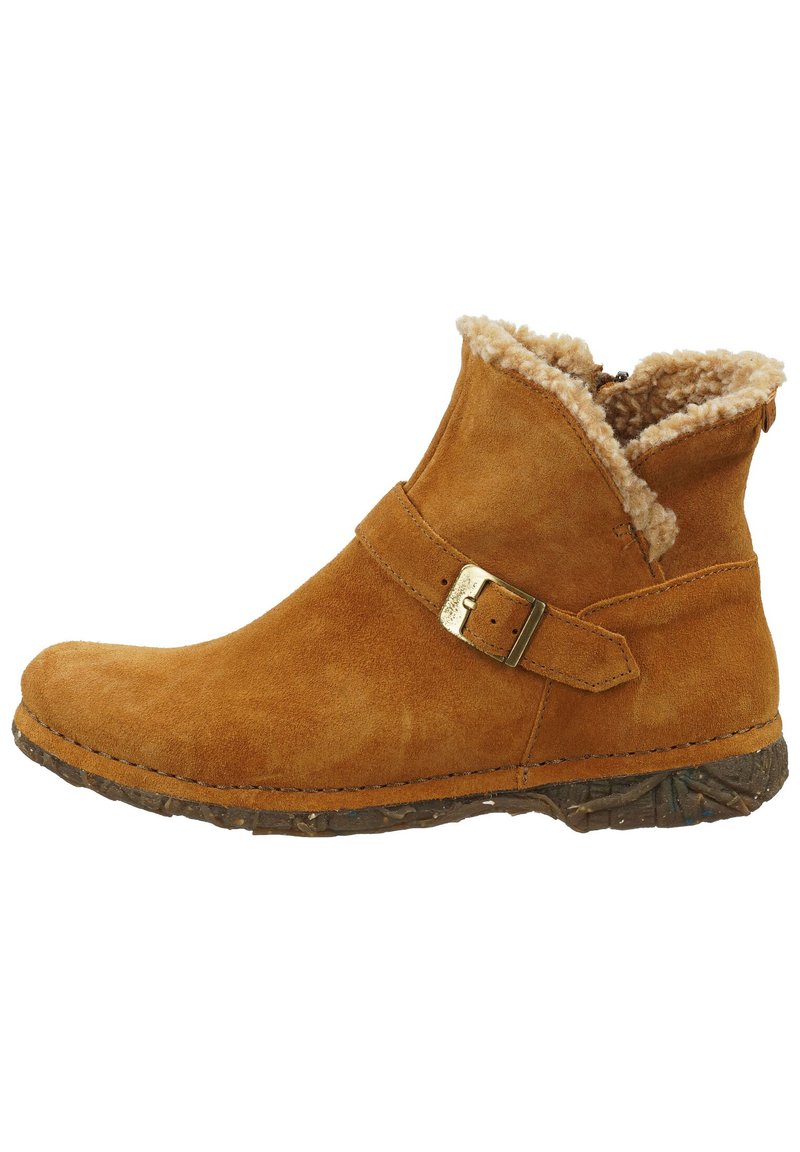 Bota de ante color marrón claro hasta el tobillo con un collar de pelo y una hebilla lateral. Suela gruesa con textura que proporciona agarre. Presenta un diseño de punta redondeada.