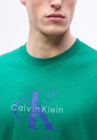 Νέος άνδρας που φοράει ένα φωτεινό πράσινο μπλουζάκι Calvin Klein με μπλε και λευκό τυπωμένο λογότυπο στο στήθος, εμφανιζόμενος από τον λαιμό και κάτω.