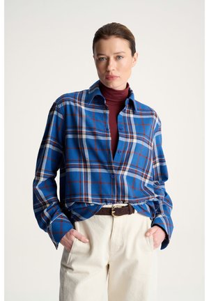 Kobieta w niebiesko-czerwoną kratę koszulę na bordowym golfie, kremowe spodnie i brązowy pasek, stoi z rękami w kieszeniach na białym tle.