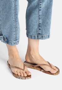 Brown flip-flops with a textured leopard print sole and thin, transparent straps. Worn with cuffed light blue denim pants.