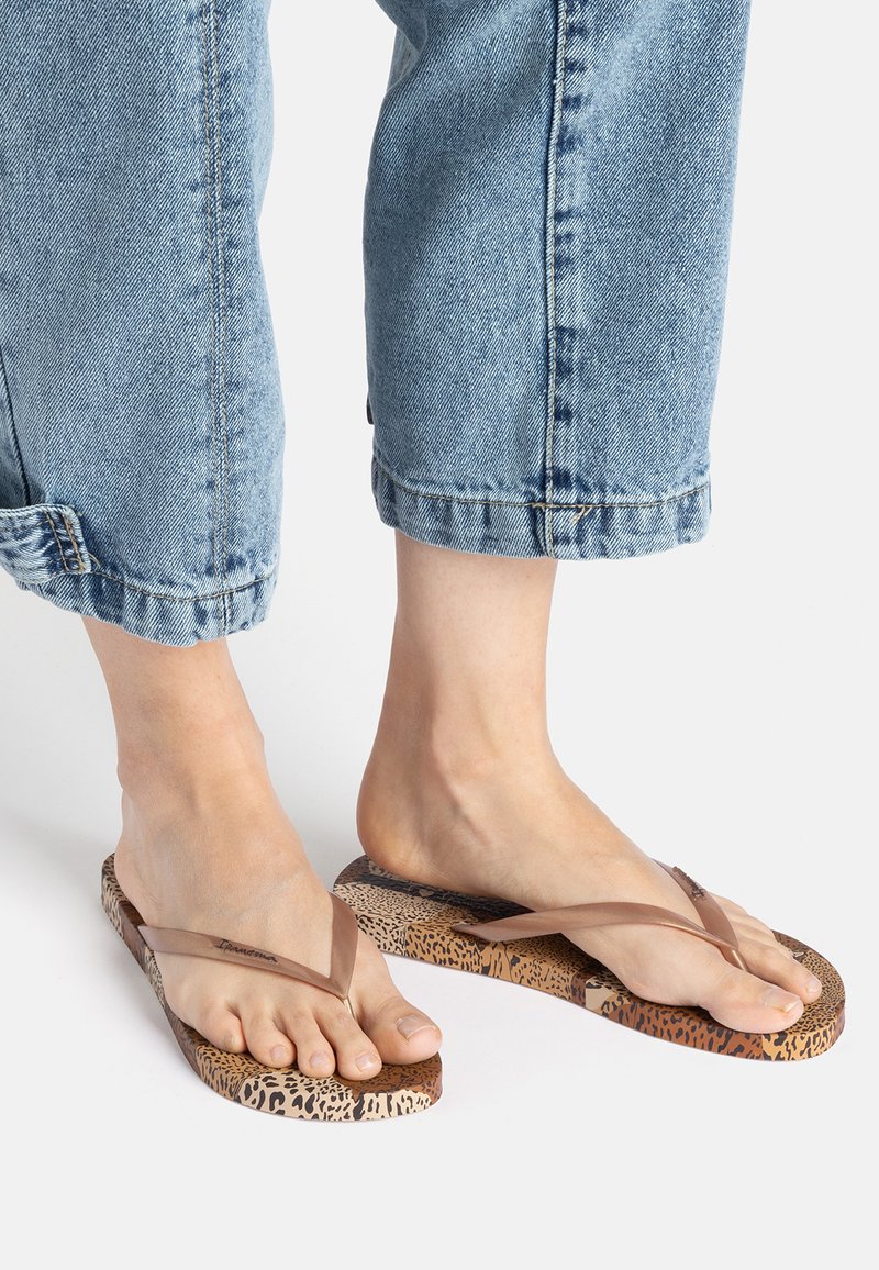 Brown flip-flops with a textured leopard print sole and thin, transparent straps. Worn with cuffed light blue denim pants.