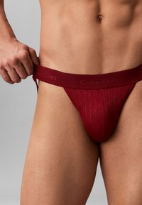 Rode Calvin Klein string, met een gestructureerd patroon en een brede tailleband met een gecursiveerd logo. Het materiaal lijkt zacht en rekbaar.
