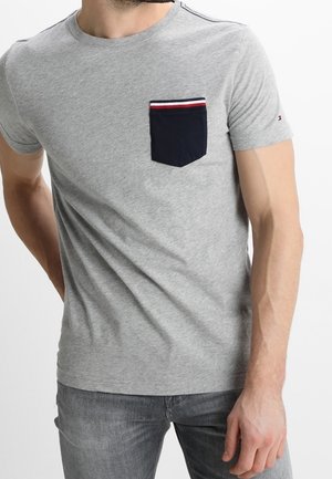 Man draagt een lichtgrijs T-shirt met korte mouwen met een zwart borstzakje voorzien van rode en witte strepen aan de bovenkant, gecombineerd met grijze jeans.