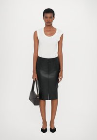 Black leather pencil skirt, fitted design with front slit, paired with a white scoop-neck tank top. Model holds a dark handbag.