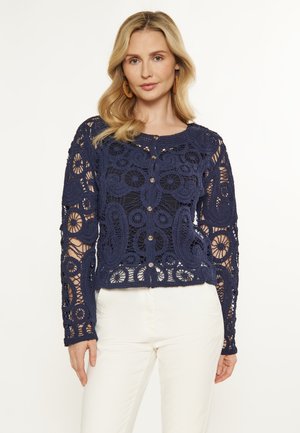 Marineblauer Häkel-Cardigan mit floralen und paisley-muster, langen Ärmeln, rundem Hals und Knopfverschluss vorne. Über hellfarbigen Hosen getragen.