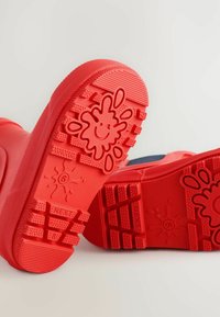 Czerwone gumowe kalosze z teksturowaną podeszwą, na której znajduje się uśmiechnięta buźka oraz wzór w plamy. Projekt zawiera głębokie rowki dla lepszej przyczepności.