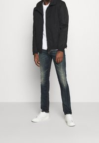 Černá zipová bunda s kapucí, bílý tričko, opotřebované tmavé denimové džíny a bílé tenisky. Neformální outfit s minimalistickým designem.