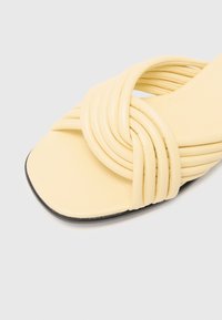 Sandal de tiras amarillas que presenta un diseño de tiras anchas y retorcidas, suela plana y sutiles acentos negros en el borde. Acabado suave y mate.