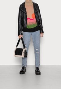 Schwarze Leder-Motorradjacke über einem beigen Pullover mit buntem Landschaftsdesign, kombiniert mit hellblauen, zerrissenen Jeans und schwarzen Plateausandalen, während eine schwarz-creamfarbene Handtasche getragen wird.