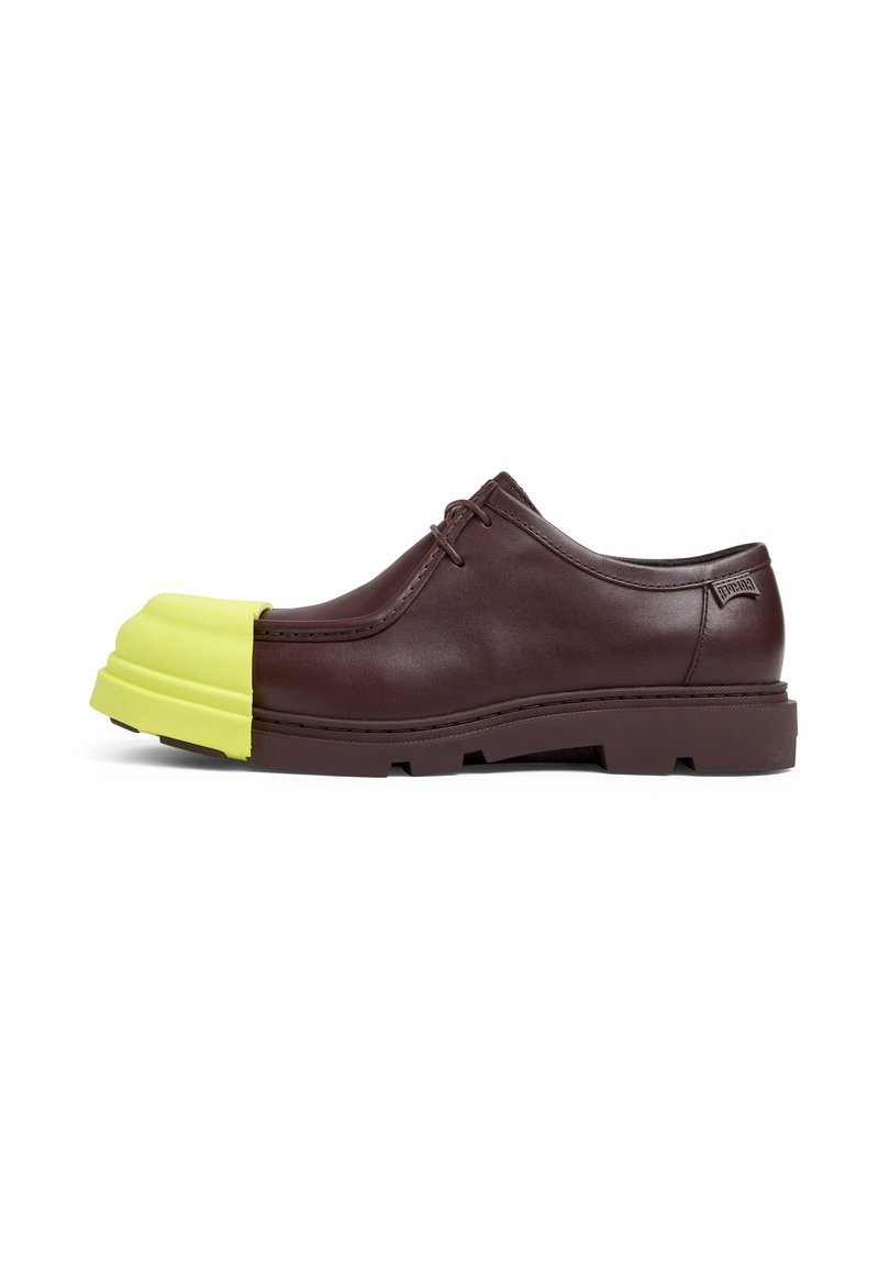 Scarpa di pelle marrone con una punta in gomma gialla brillante, chiusura con lacci e una suola spessa con un motivo testurizzato.