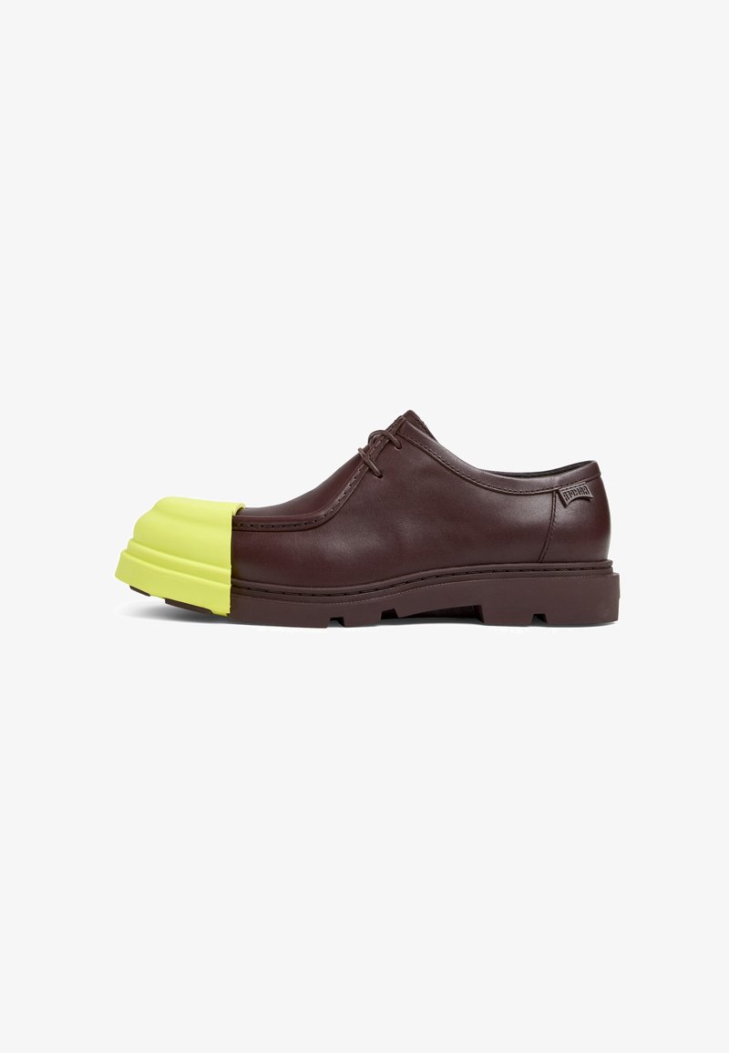 Scarpa di pelle marrone con una punta in gomma gialla brillante, chiusura con lacci e una suola spessa con un motivo testurizzato.