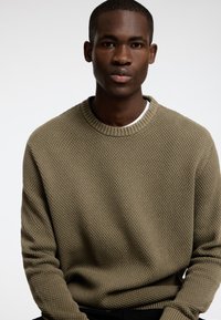 Olīvu zaļš ar tekstūru adīts džemperis ar apaļu ribotu apkakli un garām piedurknēm, apvienots ar baltu krekliņu.