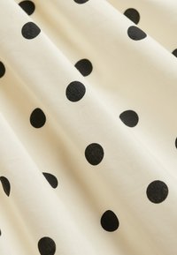 Bomuldsstof med en cremet baggrund og sorte cirkulære prikker i forskellige størrelser, der viser en glat, tekstureret overflade.