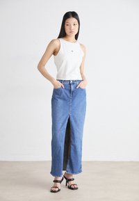 Hvit ermeløs topp parret med et høyt liv, langt denimskjørt med en frontsplit, medium blåfarge og rett silhuett. Sorte høyhælte sandaler.